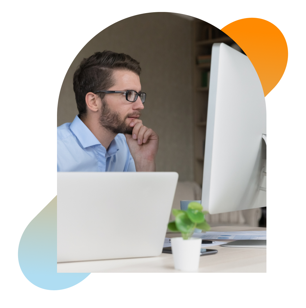 Contemplative man wearing glasses seated in front of computer, staring at large monitor.